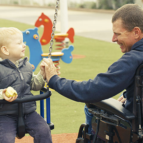 Mann im Rollstuhl spielt mit seinem Sohn auf Spielplatz. 